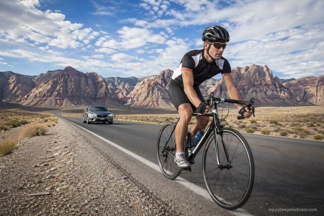 Cyclist riding on a desert highway with mountains and a car behind; small “injurylawyeradvisor.com” text in the corner.