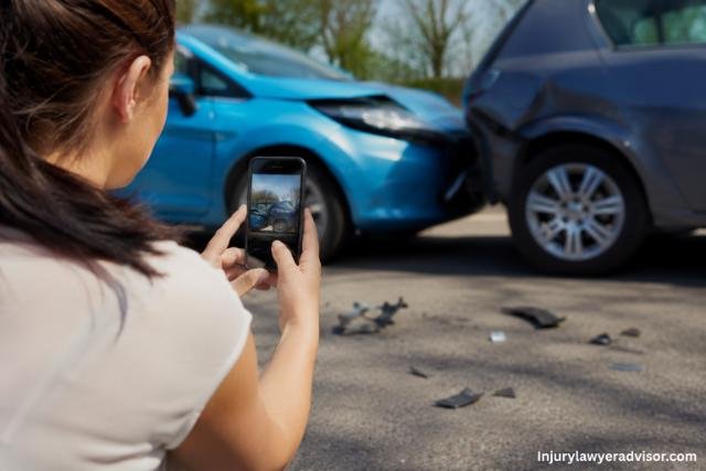 A woman photographs a car accident scene with her smartphone while damaged vehicles and debris are visible on the road in the background.